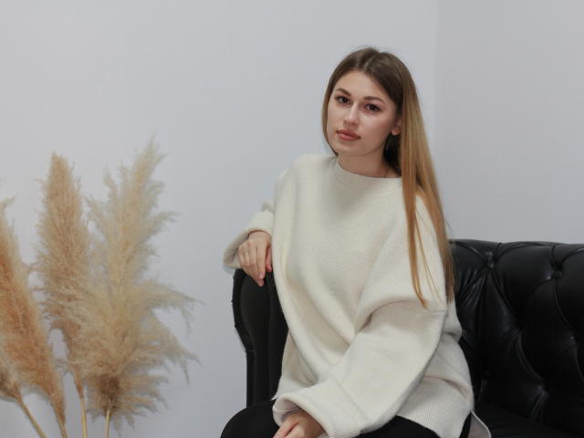 AlinaNathan with long brown hair, wearing a white chunky sweater, sitting on a black leather couch, hands on her knee, pensive expression, pale skin, and soft pink lipstick.