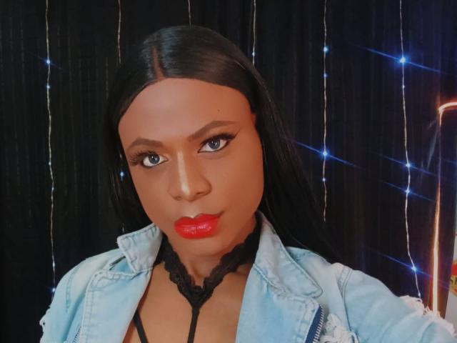 VenusMiller has a medium brown skin tone and long, straight black hair. She's wearing a light blue denim jacket with a black lace choker. Her red lipstick contrasts sharply with her black eyeliner and eye makeup. The image focuses on her face, which is centered, with a slight upward angle. She has a confident, direct gaze. The background features a black curtain with string lights, adding a subtle, festive glow. The composition is tight, emphasizing her facial features and attire. The photo has a slightly warm tone.