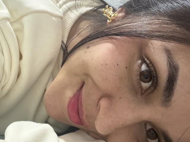 HikariChloe's face, brown eyes, pink lips, white sweater, small freckles, black hair with gold hair clip, lying down, close-up shot, slightly smiling.