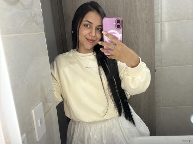 HikariChloe with long black hair, smiling, holding a pink phone, wearing a white sweatshirt and white skirt, standing in a bathroom, showing her face and upper body.
