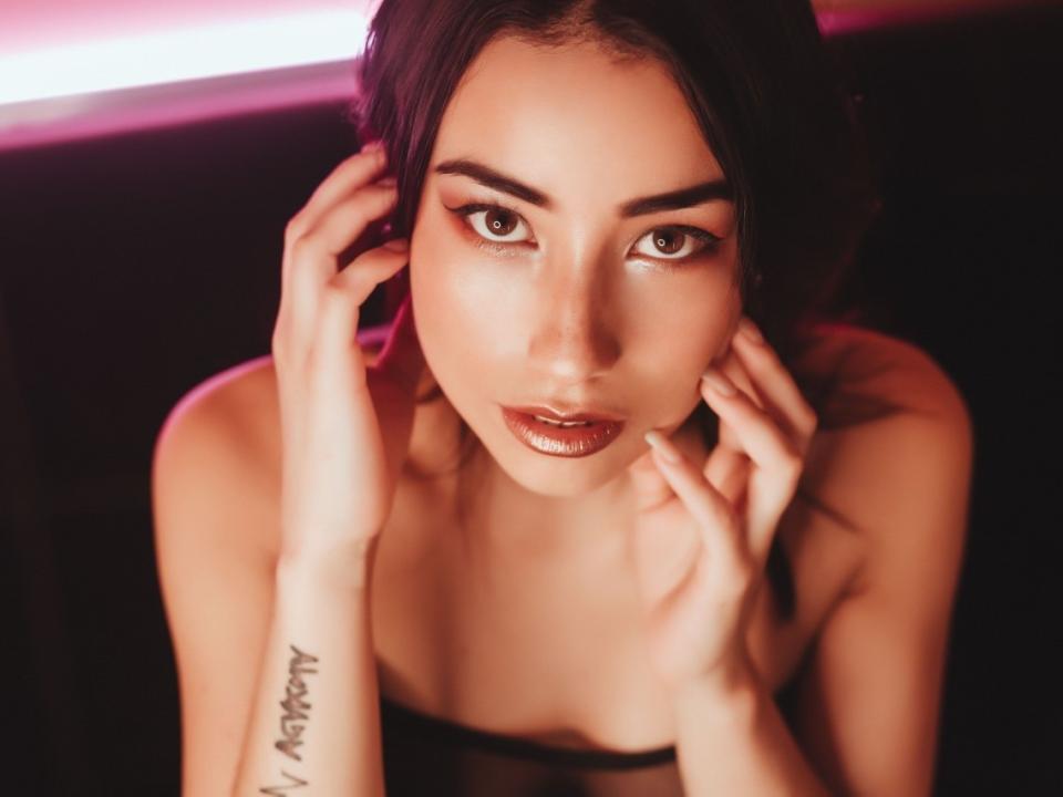 Valeryy69 has shiny brown eyes, dark hair, and is touching her face with hands, showing a tattoo on her left wrist. She has glossy lips and is looking up.