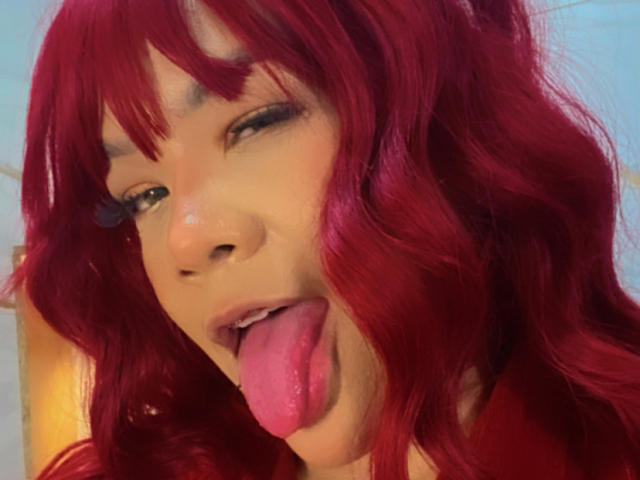 Close-up of VictoriaField with bright red, wavy hair and bangs. She's sticking out her pink tongue, slightly wrinkled. Her eyes are half-closed with long, dark eyelashes. She has a light brown skin tone. The lighting is warm, highlighting her hair and face. The background is blurred, focusing on VictoriaField's facial expression and hair texture. The image uses a digital effect to emphasize her hair's vibrant color.