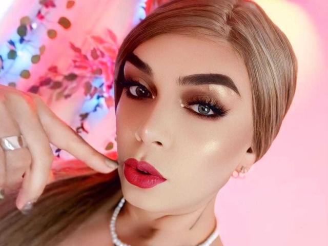 KarolElegance tiene un lápiz labial rojo rosado brillante, pestañas largas y oscuras, y está señalando su rostro con un dedo. Lleva un collar blanco y tiene un anillo de plata..