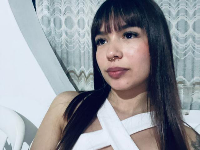Eileen Swift ha i capelli castano lisci con la frangia, un piercing al naso e indossa un top bianco a fascia che mette in mostra un seno piccolo e un sorriso appena accennato. La sua pelle è chiara ed è carina..