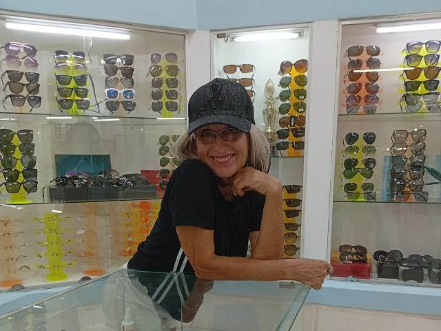 ElizaWendy with gray hair, black cap, and glasses, smiling at the camera, leaning on a glass counter, surrounded by multiple sunglasses.