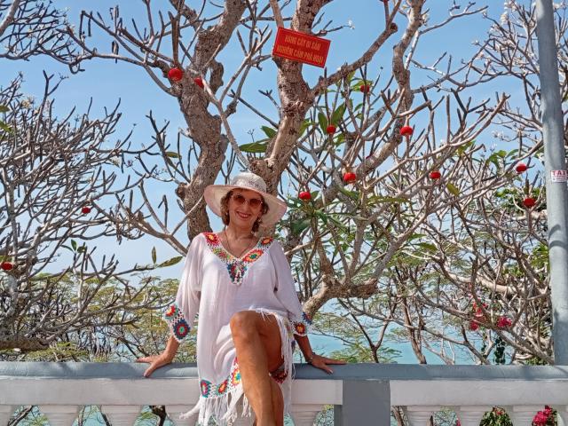 ElizaWendy with a white dress, colorful embroidery, and a sunhat, sitting on a blue railing, tree with red apples behind, bright blue sky, and a clear ocean in background.