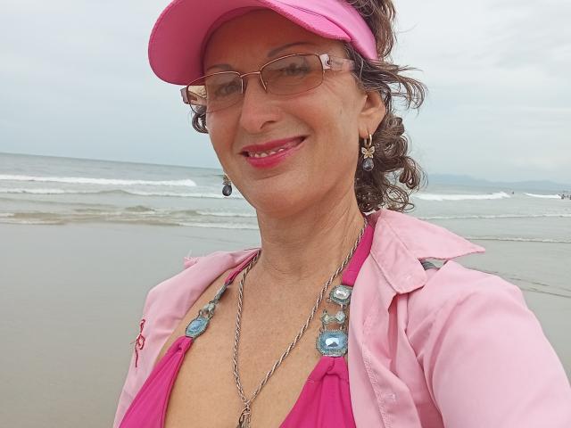 ElizaWendy, middle-aged woman with pink visor, glasses, curly brown hair, pink bikini top, pink jacket, blue necklace, smiling, pink lipstick, beach background.