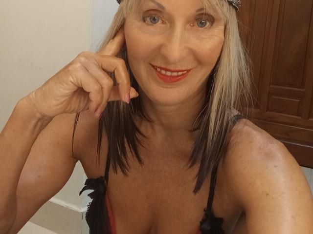 ElizaWendy has blonde-black dyed hair, red lipstick, black lace bra, smiling, touching her ear, showing her tits, and wearing a black headband in a selfie.