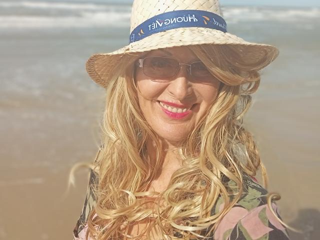 ElizaWendy with blonde wavy hair, wearing a straw hat with a blue band, sunglasses, and pink lipstick, smiling at the beach, in a camo-patterned shirt.