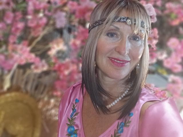 ElizaWendy with pink dress, flower headband, pearl necklace, and blue embroidery, has a white smudge on her face, smiling with pink lipstick, and brown blonde hair.