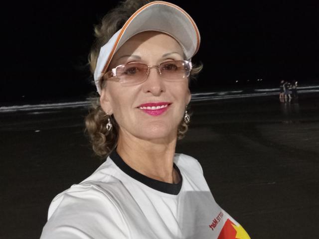 ElizaWendy with curly brown hair, glasses, white visor, pink lipstick, and white shirt, smiling at the camera, night beach setting, visible sand and small group in background.