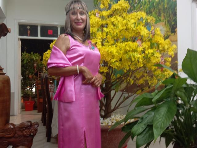 ElizaWendy, middle-aged woman with gray hair, pink dress, and exposed shoulder, stands smiling, showing her tits, with yellow flowers in the background and green plant on the right.
