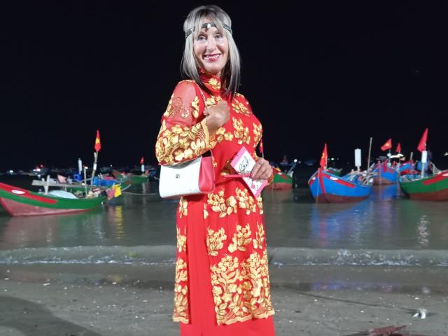 ElizaWendy, Asian woman, red and gold floral dress, smiling, holding white purse, standing on beach, colorful boats in background, night, blonde hair with bangs, medium build, cute.