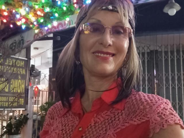 ElizaWendy has medium-length brown hair, wears a red crocheted shirt, and pink glasses, smiling with red lipstick, decorated with colorful Christmas lights in the background.