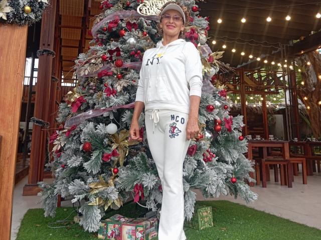 ElizaWendy stands in front of a decorated Christmas tree, wearing a white hoodie and pants with a "No Po" patch, smiling with her hands by her sides.