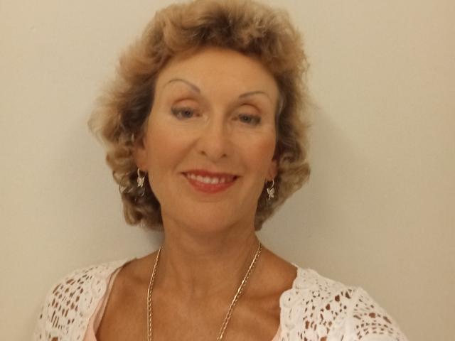 ElizaWendy has curly blonde hair, red lipstick, and wears a white lace top with earrings and a gold necklace, smiling gently. Her tits are small, and she's attractive.