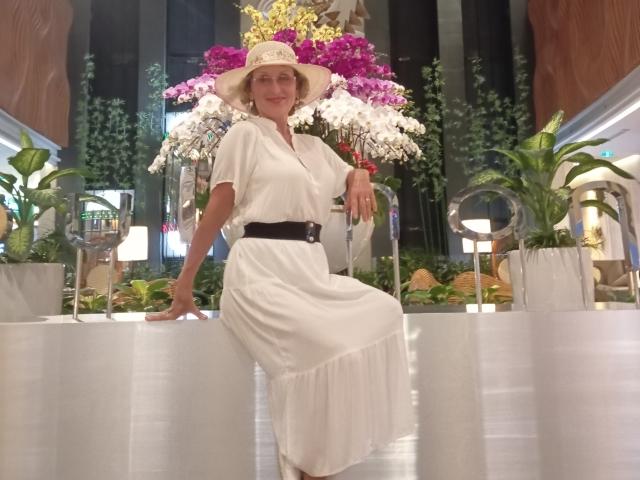 ElizaWendy is a sexy middle-aged woman with a white dress, black belt, and wide-brimmed hat, sitting on a white table, surrounded by colorful flowers and green plants.