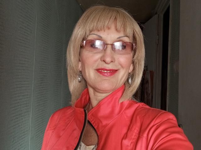 ElizaWendy has blonde bob, pink glasses, red lipstick, and a shiny red jacket with a zipper. She's smiling, showing her teeth, and has silver earrings.