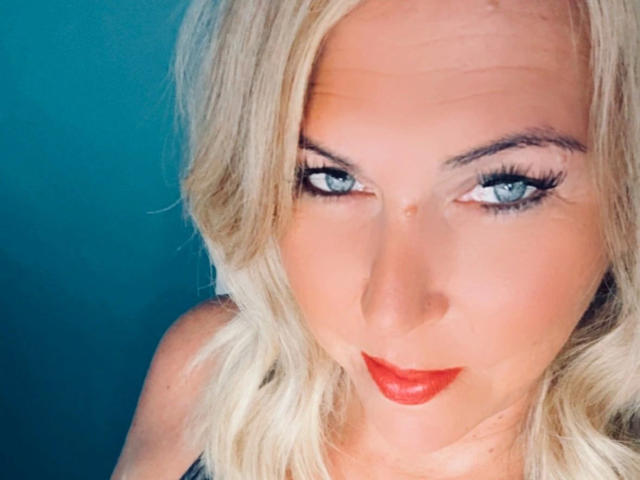 AurelieNoir-hot tiene cabello rubio, ojos azules y lápiz labial rojo. Sus senos son ligeramente visibles en el encuadre. Está mirando hacia arriba con una expresión sexy y segura..
