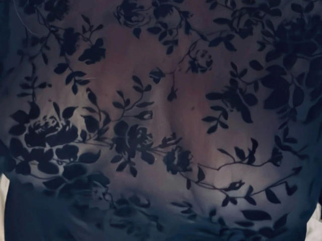 AurelieNoir-hot's chest, covered in a sheer black floral-patterned top, showing subtle, soft-breast outlines and a slightly visible, rounded belly underneath.