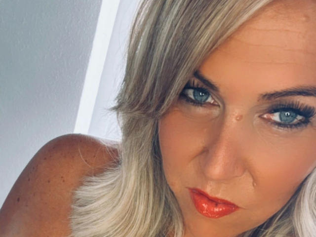 AurelieNoir-hot tiene cabello rubio, ojos azules y labios rojos brillantes, con un pequeño piercing en la nariz, mirando fijamente a la cámara con una expresión seria y con algunas pecas visibles..
