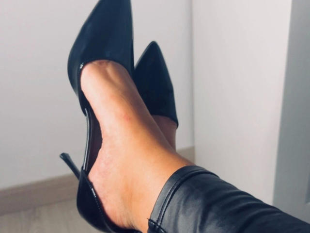 De benen van AurelieNoir-hot, gekleed in zwarte, puntige, hoge laarzen, met haar rechterbeen over haar linkerbeen gekruist, waardoor haar glanzende, met leer beklede kuiten zichtbaar zijn..