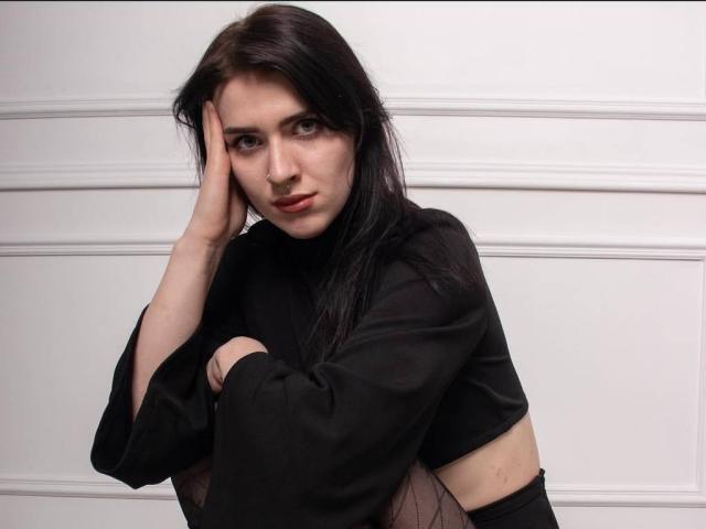 DarinaIngrid with dark, straight hair, wearing a black outfit, has her hand on her head, looking serious, with a slight frown, and her arm crossed over her chest.