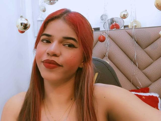KairaDayamai has red-dyed straight hair, full lips with dark red lipstick, and a cute smile. She's topless, showing her bare shoulders and a gold necklace.