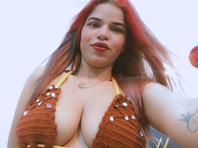 KairaDayamai with red hair, wearing a brown crochet bikini top showing her big boobs, yellow straps, nose ring, and tattoo, smiling slightly, looking down at the camera.