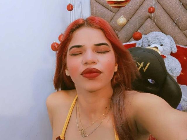 KairaDayamai with red hair, closed eyes, and red lipstick, wearing a yellow top, necklace, and gold pendant, smiling, with a stuffed bear and hat in the background.