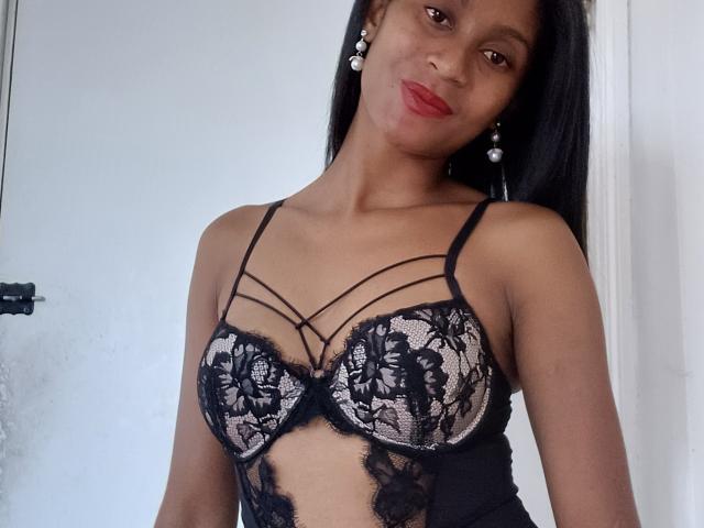 PerlaLouisa with dark skin, long black hair, wearing black lace bra with floral patterns, strappy design, showing her tits, red lipstick, pearl earrings, white wall background.
