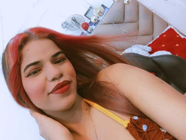 KairaDayamai with red and brown hair, yellow top, red lipstick, smiling, touching her head, in a cozy room, showing her tits slightly visible in the brown top.