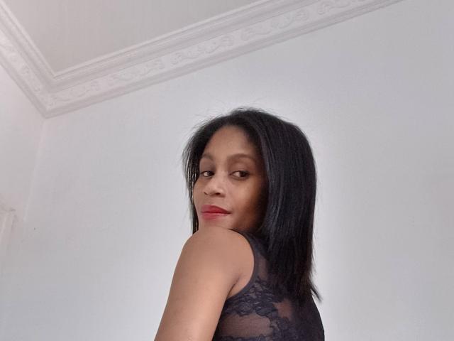 PerlaLouisa with medium brown skin, straight black hair, red lipstick, and a black lace sleeveless top, looking over her shoulder, showing her bare back and small ass.