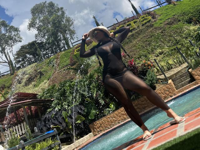 AlahiaRouse stands in a pool, wearing a sheer black dress that reveals her slim, toned physique and small breasts. She has dark skin and is wearing a wide-brimmed hat. Her right leg is bent and her left arm is raised, holding the hat. The pool water is clear and blue. She is barefoot on a red-tiled pool deck. Greenery and a wooden structure are in the background. The image is brightly lit with natural sunlight.