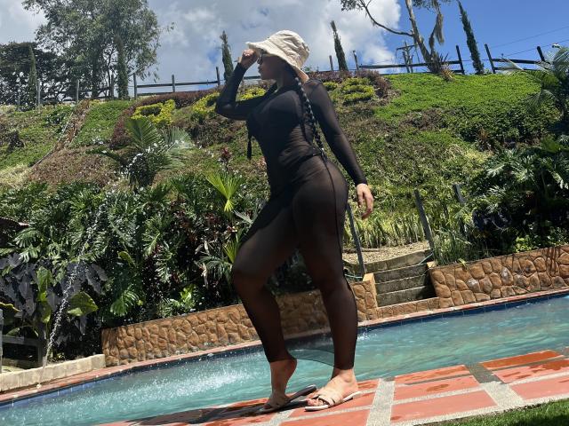 AlahiaRouse stands near a pool, wearing a sheer black long-sleeve bodysuit and beige bucket hat. She's looking up with her right hand shading her eyes. Her full, dark brown legs are visible through the bodysuit. She's barefoot with beige sandals on. The pool has clear blue water and a stone edge. The background features lush greenery and a hill with trees and a wooden fence. The sky is blue with scattered clouds. Her body is curvy with a large butt.