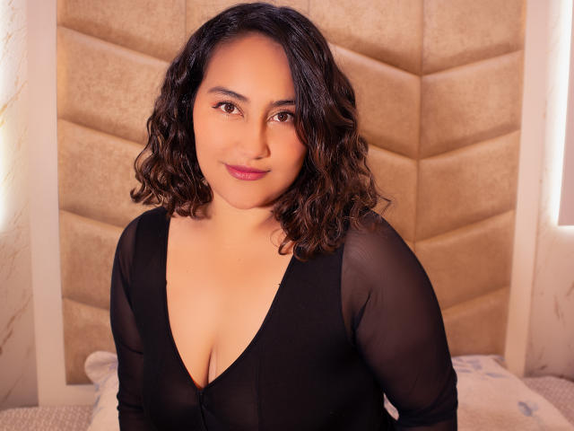 CelesteAdler with curly black hair, medium brown skin, and a cute smile, wearing a sheer black top with a deep cleavage, sitting in front of a padded headboard.