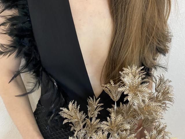 Amberia's back, showing a deep black dress with feathery sleeves, holding gold decorative branches near her tits, long brown hair falling over her shoulder.