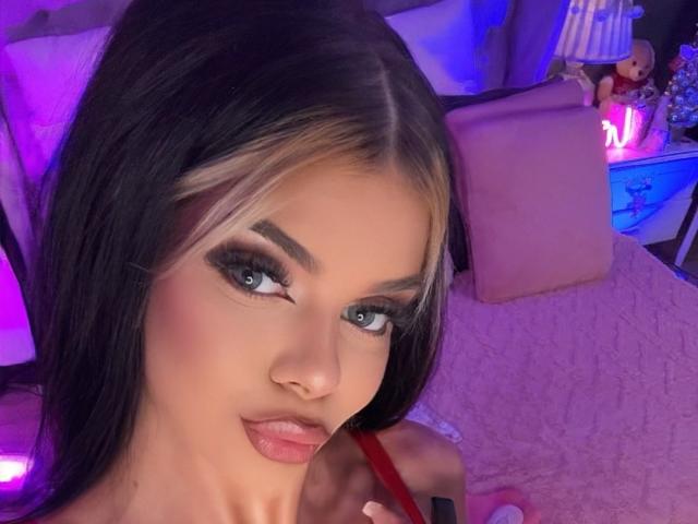 IvyRaven has long black and blonde hair, blue eyes, and full pink lips with glossy lipstick, holding a red strap in a purple-lit room.