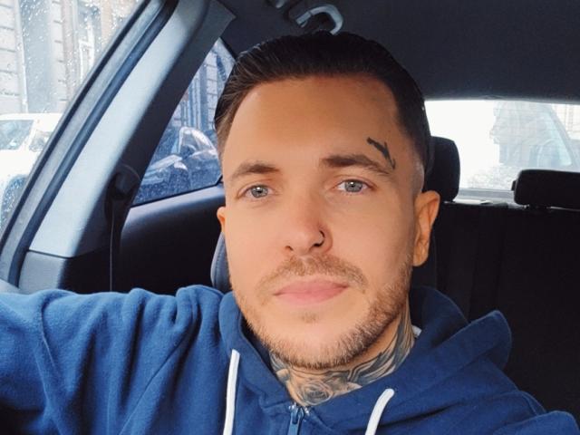 VixenMan has a clean-shaven face with a small black tattoo above his right eyebrow, blue hoodie, tattoos on his neck, short brown hair, and a slight mustache.