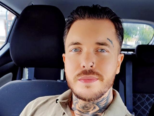 VixenMan with short dark hair, blue eyes, beard, nose ring, tattoo on left eyebrow, tattooed neck, smiling, wearing beige shirt, sitting in car, visible black headrest.