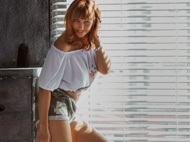 LadyNataly-hot with red hair, off-shoulder white top, and green shorts, standing by a window, sunlight casting stripes, smiling, hand near neck, looking sexy, slim, cute.