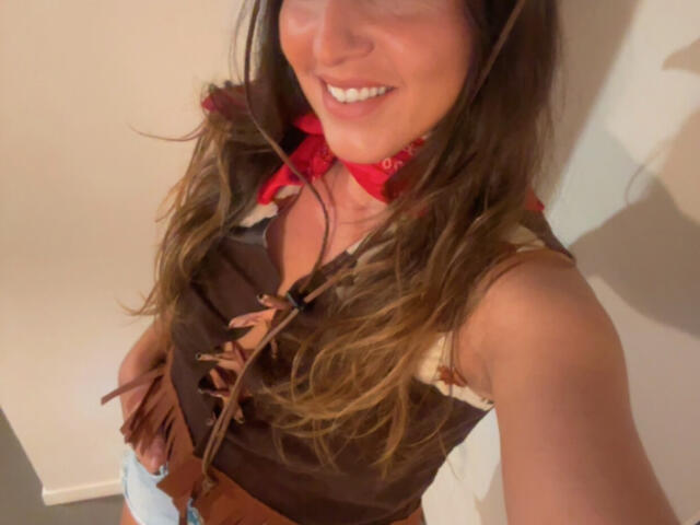 ChiaraSexyCul has long brown hair, a smiling face, and wears a brown cowboy top with lace-up front, red bandana, and fringed sleeves, showing her tits and small ass.