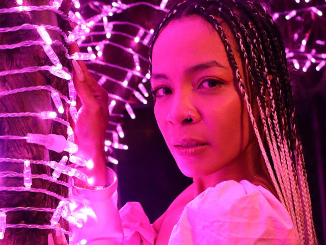 NilaStorm with braided hair, pink nose ring, and white shirt, holding a tree wrapped in pink lights. Her face is illuminated by the neon pink glow.