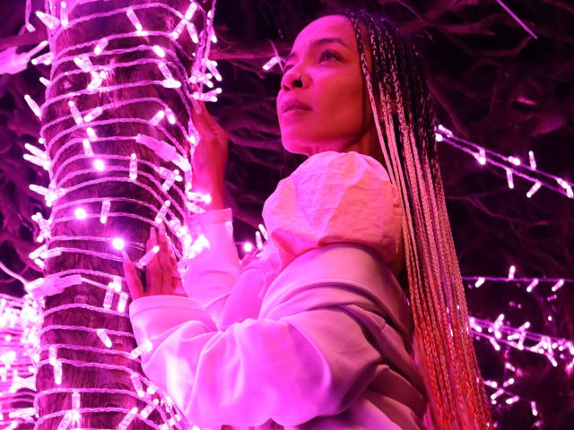 NilaStorm with long blonde braids, wearing a white puffy jacket, standing next to a tree wrapped in pink Christmas lights, looking up with a slightly pouty expression.