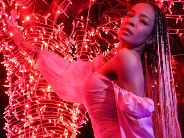 NilaStorm with long braided hair, wearing a pink off-shoulder dress, touching red string lights, looking back with a slight smile, red lighting on her brown skin.