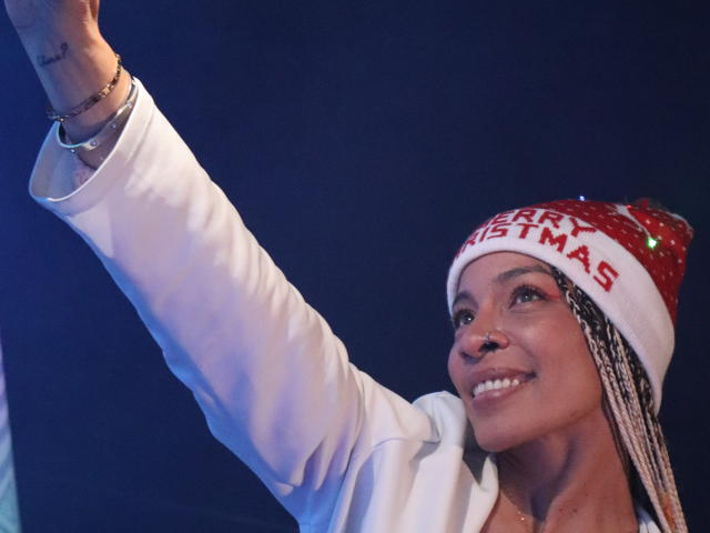 NilaStorm with dark skin, wearing a red and white Christmas hat, smiling, raising her right arm. She's in a white jacket with braids and a nose ring.