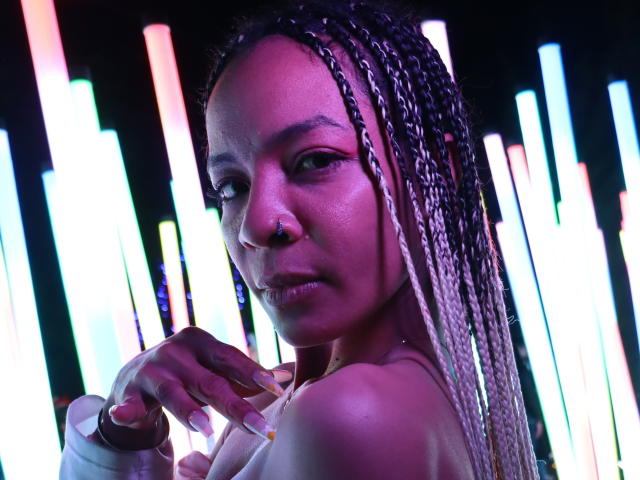 NilaStorm with dark skin and braided pink and white hair, nose ring, and yellow nails, looking at camera with a slight smirk, showing her bare shoulders and upper chest.
