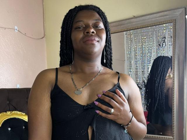 BlackVelvets with dark skin and braided hair, wearing a black lace dress, has purple nails, hand on chest, smiling, heart necklace, and ring, standing in front of mirror.
