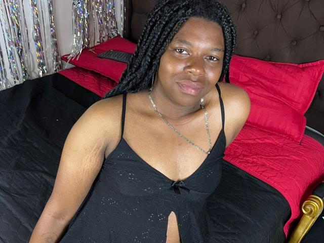 BlackVelvets, a Black woman with braided hair, small tits, and a cute smile, wearing a black dress with a keyhole, sits on a bed with red pillows.