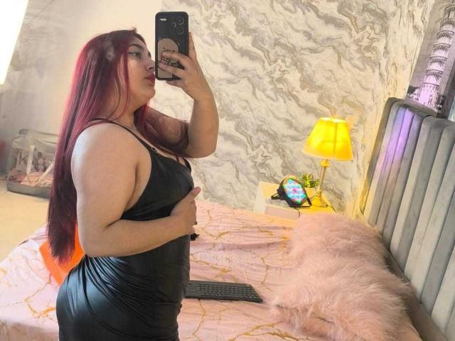 ZoeEvansl with red hair, wearing a tight black dress, taking a selfie in a bedroom. She's holding her big ass with one hand. Bed with pink and yellow decor.
